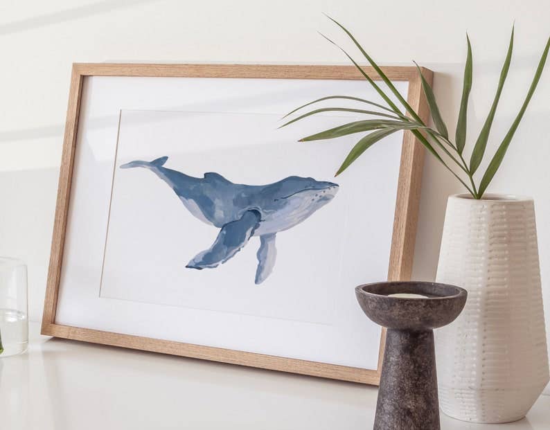 Framed whale art print with a blue watercolor illustration, styled on a white surface with a white textured vase, a dark ceramic candle holder, and a glass.