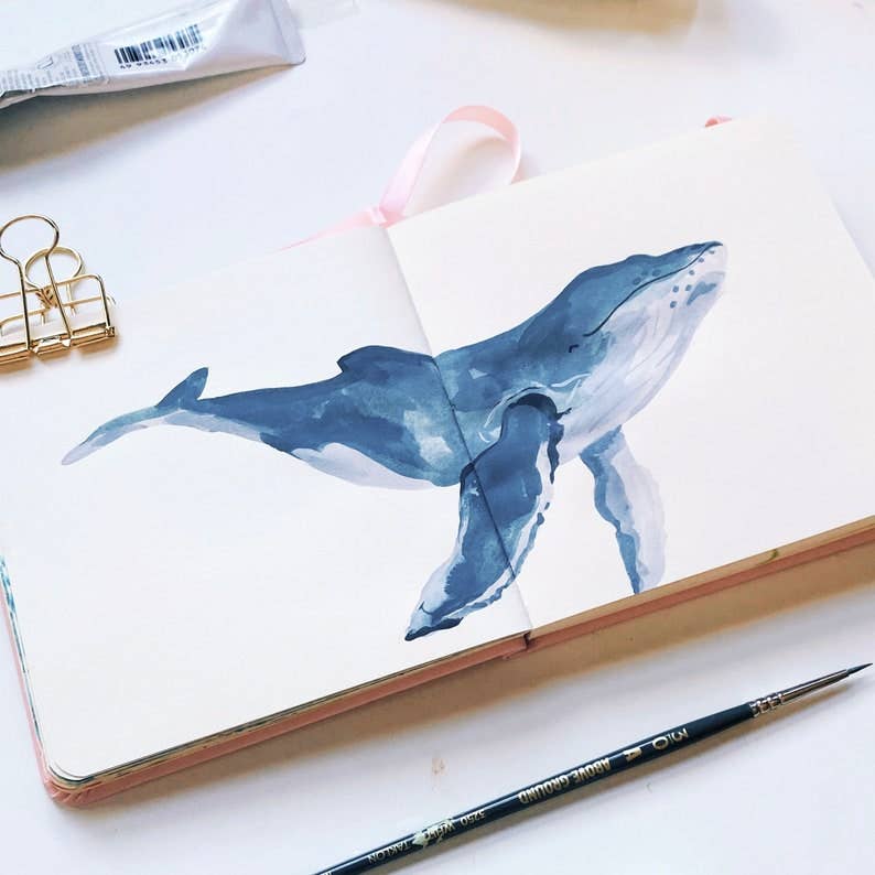 An open sketchbook with a painted blue whale on the pages, styled on a white surface with a paint tube, gold binder clips, a pink ribbon bookmark, and a paintbrush nearby.