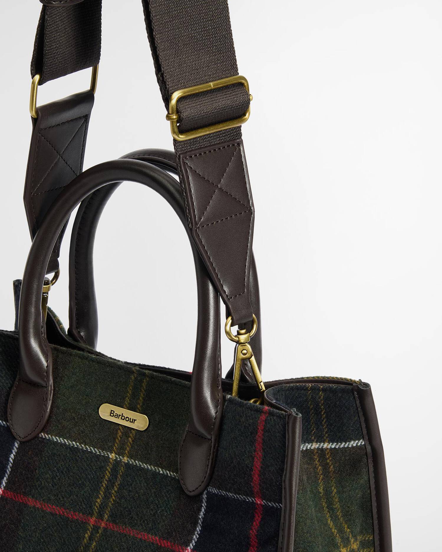 Close-up of a Barbour tartan tote bag with dark brown leather handles and a gold-tone logo plate, set against a clean white background.