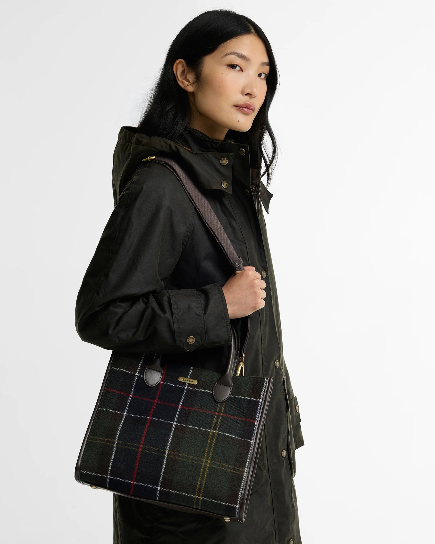 A model wearing a black hooded jacket carries a large tartan plaid tote bag with brown leather trim and gold-tone hardware against a plain white background.