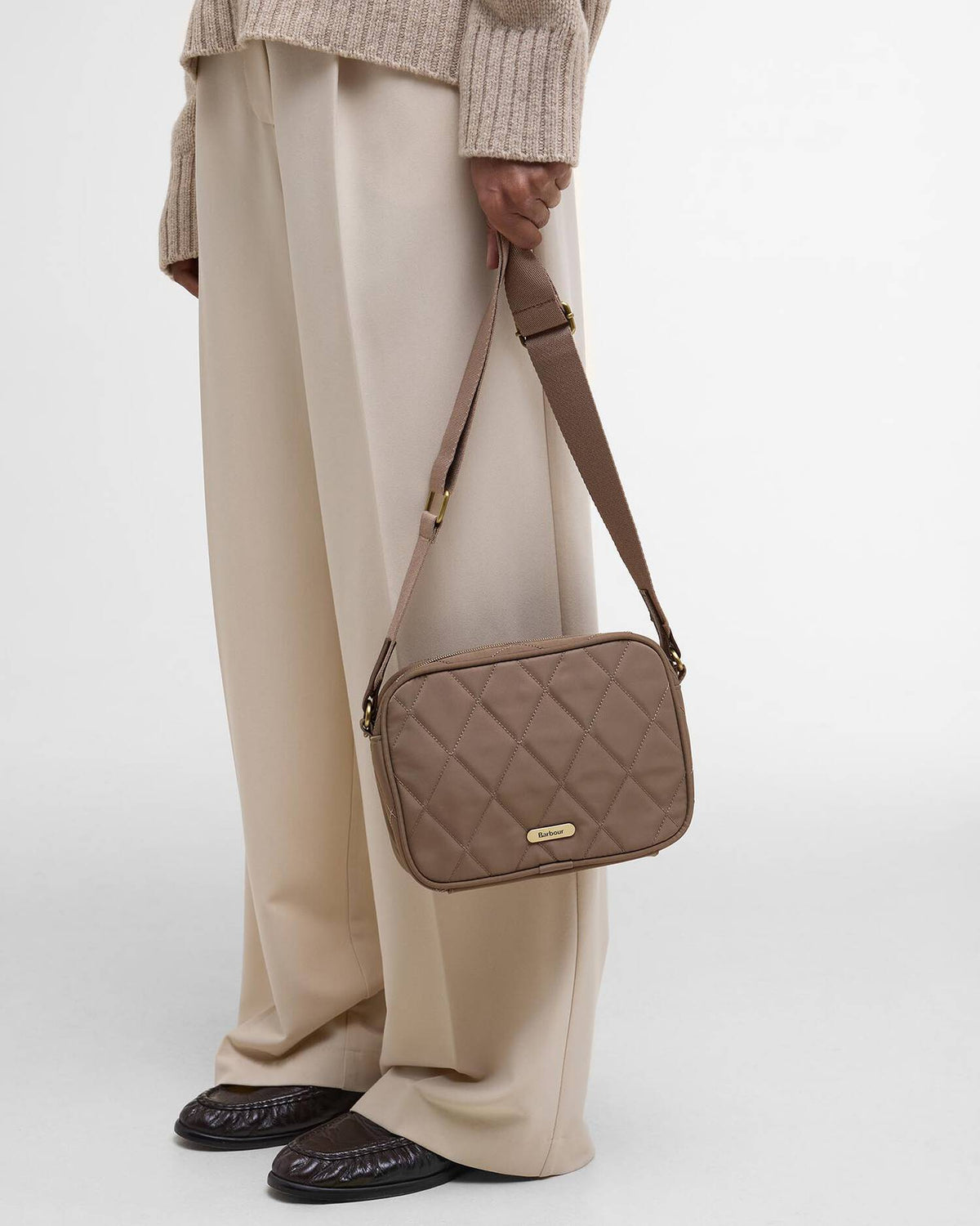A person wearing beige wide-leg trousers and a ribbed knit sweater holds a taupe quilted crossbody bag with a gold-tone logo plate. The photo is taken against a clean white studio background.