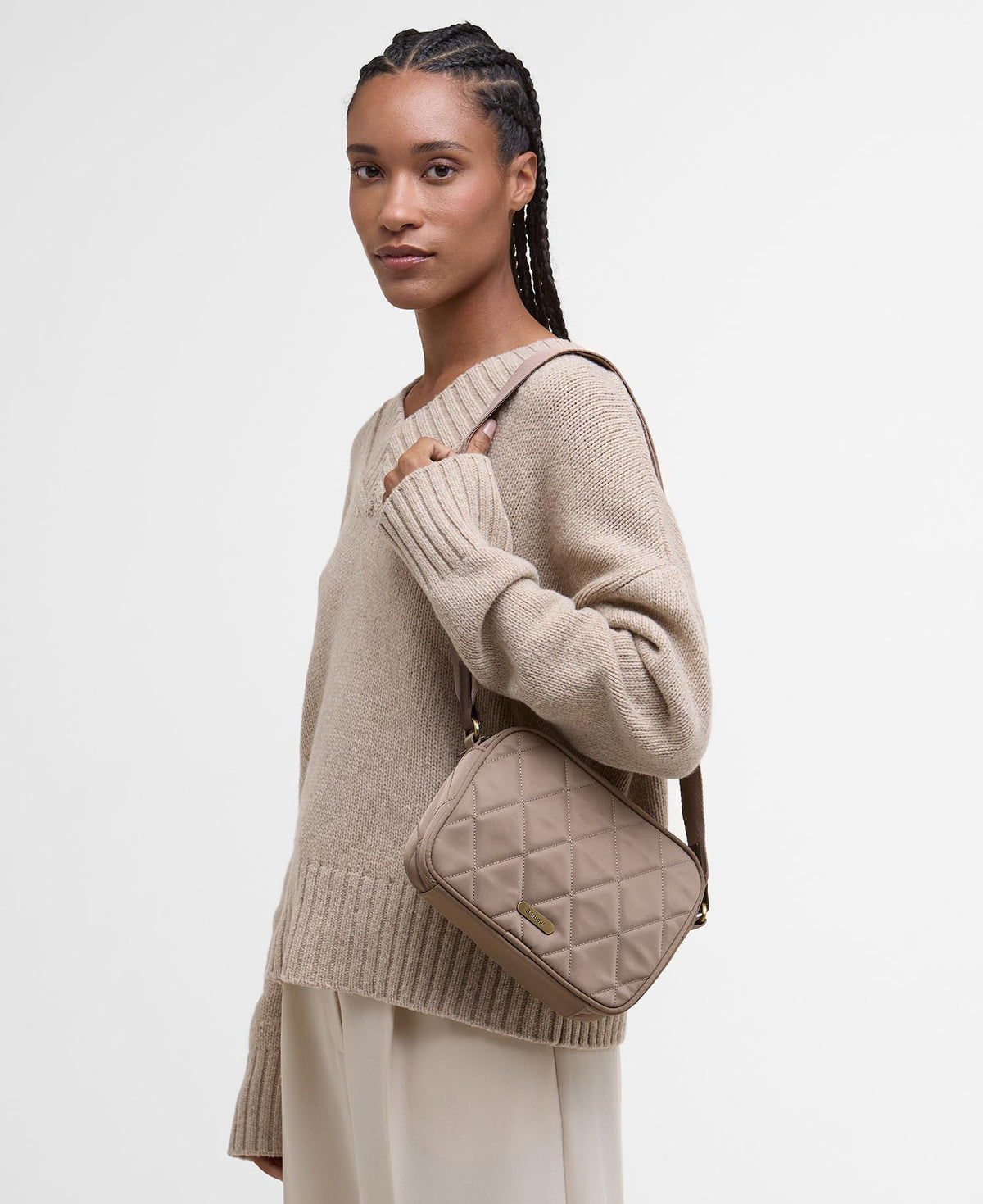 A woman wearing a beige knit sweater and cream pants carries a quilted taupe crossbody bag with a small gold logo plaque, photographed against a clean white studio background.