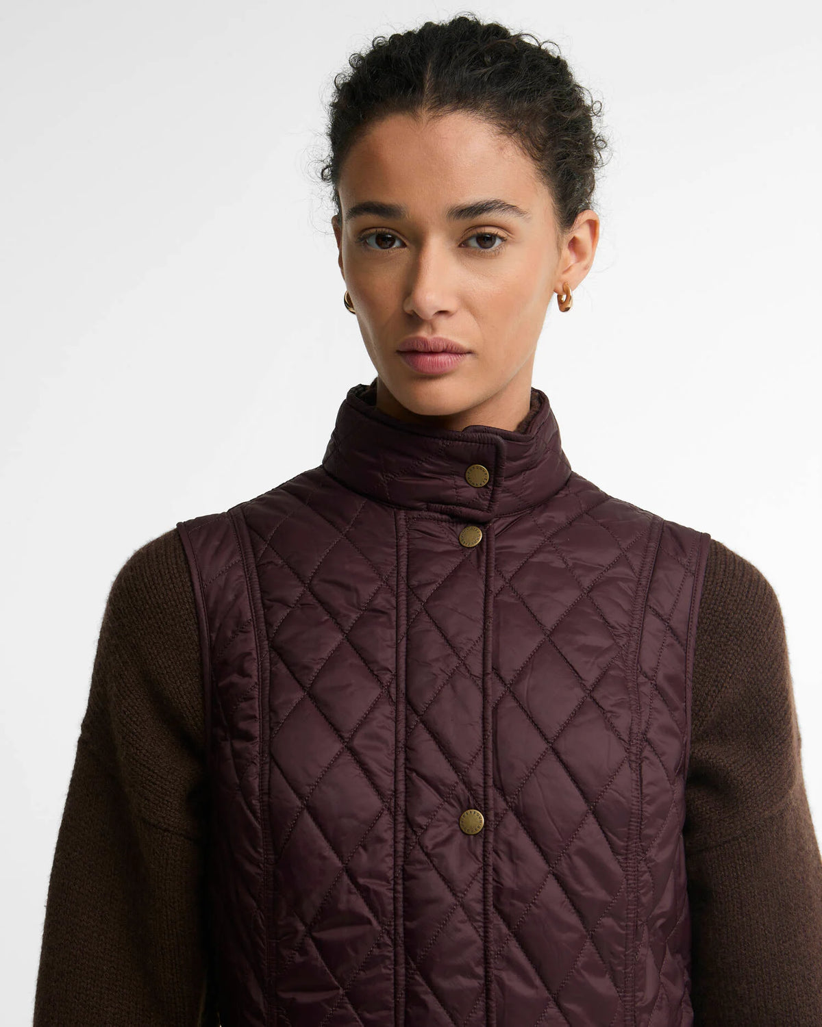 A model wearing a dark plum purple diamond-quilted puffer vest over a brown knit sweater, photographed against a clean white studio background.