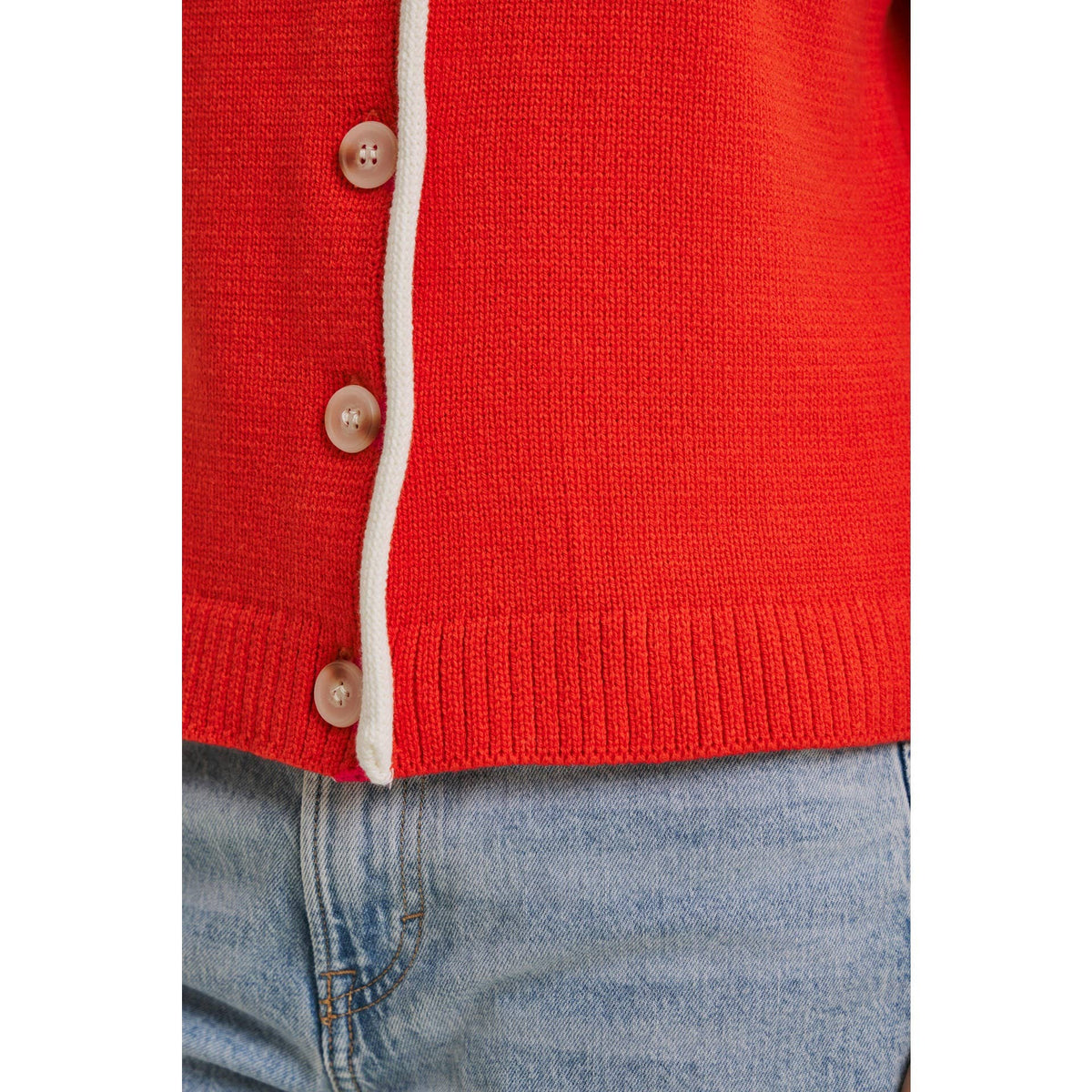 Red cardigan with white trim and brown buttons worn over blue jeans on a white background