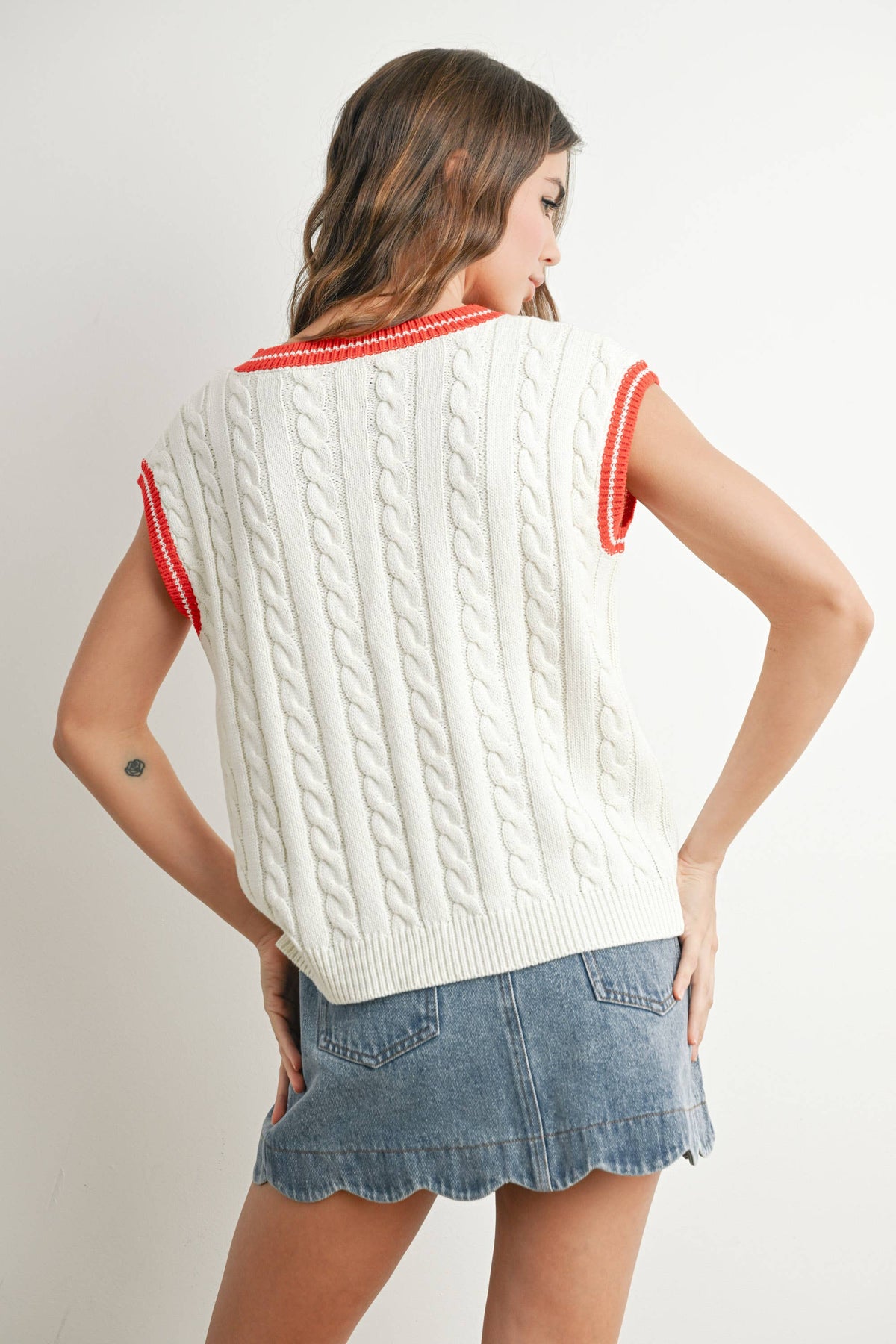 A woman shown from the back wearing a cream cable-knit sleeveless sweater vest with red trim on the collar and sleeve cuffs, paired with a denim skirt, against a light gray studio background.