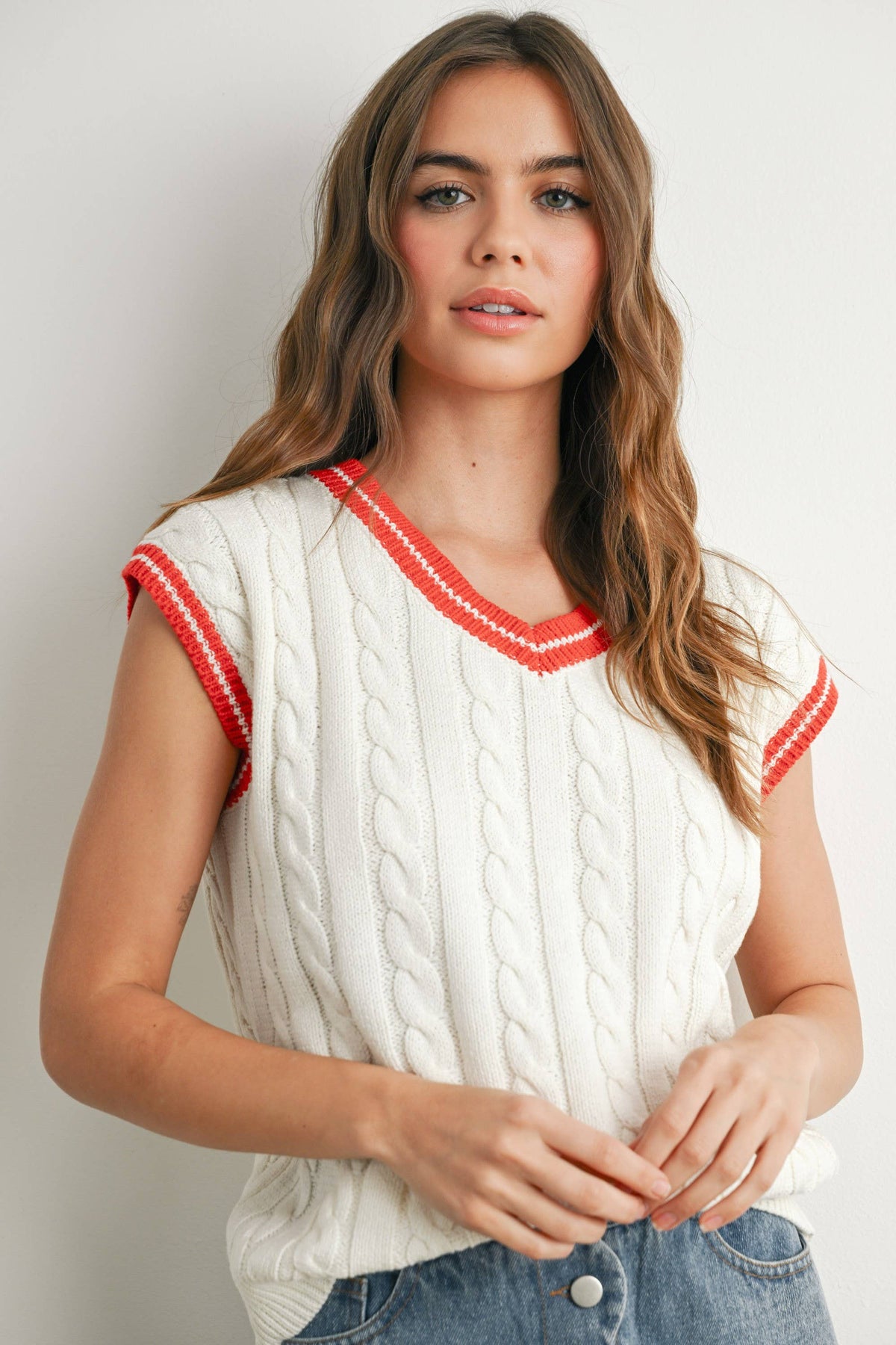 A woman wearing a cream cable-knit sleeveless sweater vest with red and white striped trim on the V-neck and sleeve cuffs, paired with blue jeans against a light gray background.