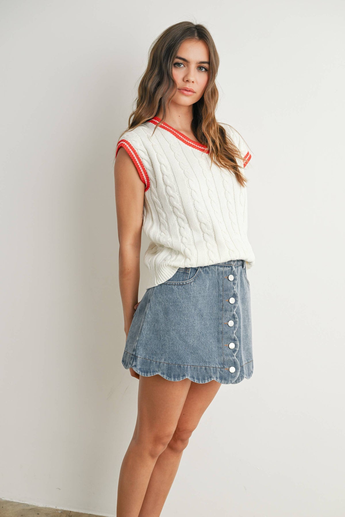 A woman modeling a white cable-knit sleeveless top with red trim and a light blue denim button-front mini skirt against a plain off-white wall.
