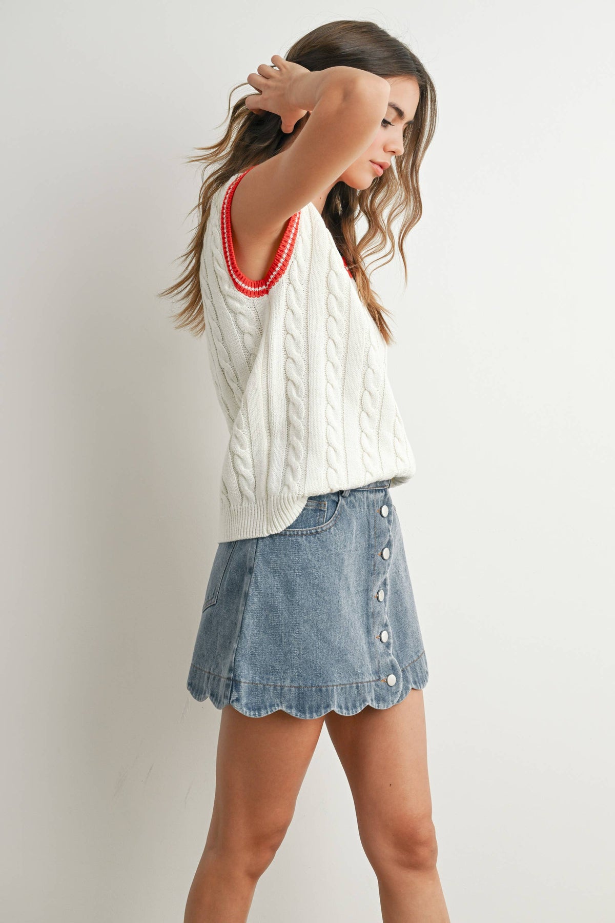 A woman modeling a white cable-knit sleeveless sweater vest with a red trim, paired with a light blue denim mini skirt with scalloped edges and button details, against a plain light gray background.
