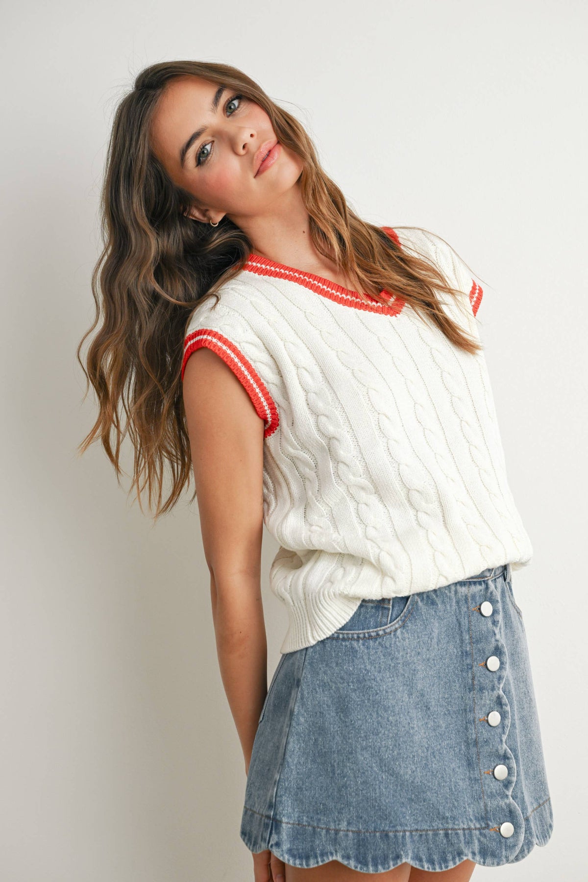 A woman wearing a white cable-knit sleeveless sweater vest with red trim and a light blue denim button-front skirt, photographed against a plain light gray studio background.