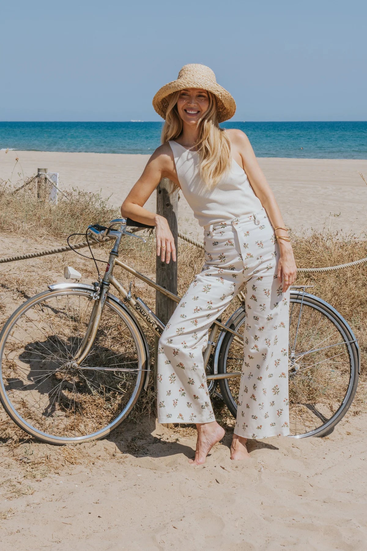 A woman stands barefoot on a sandy beach next to a bicycle, wearing a straw hat, a sleeveless cream top, and cream wide-leg pants with a small floral print. The ocean and blue sky are visible in the background.