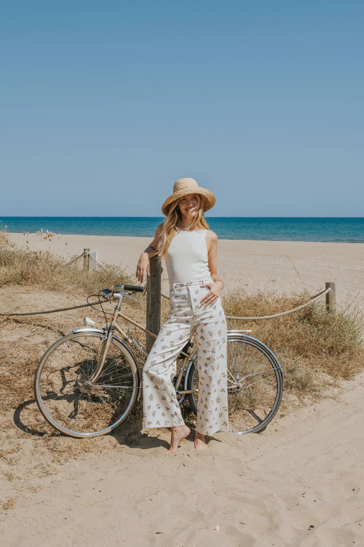 A woman stands on a sandy beach path next to a bicycle, wearing a straw hat, white sleeveless top, and white wide-leg pants with a small floral print. The background features a blue ocean and clear sky.
