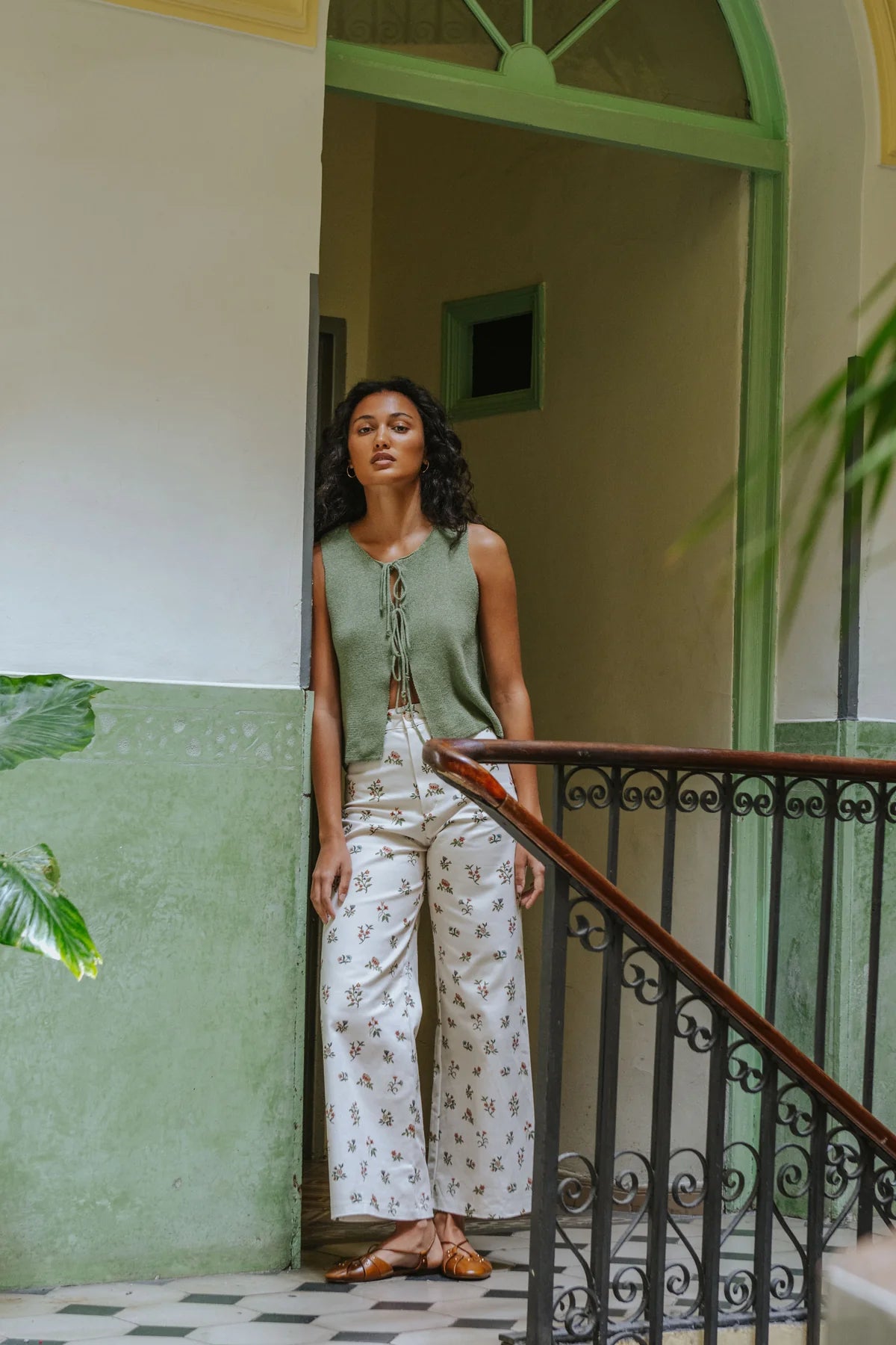 A woman stands in a doorway wearing a sage green sleeveless top with a front tie detail and white wide-leg pants with a small floral print. She is posed next to a staircase with a dark metal railing and a green-painted wall.