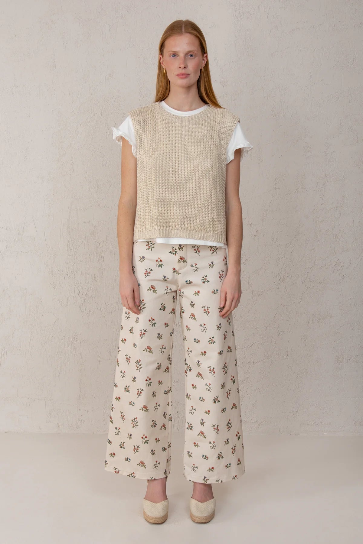 A woman stands against a textured light beige wall wearing a cream ribbed sleeveless knit top over a white ruffled short-sleeve shirt, paired with cream wide-leg pants featuring a small brown floral print, and beige pointed shoes.