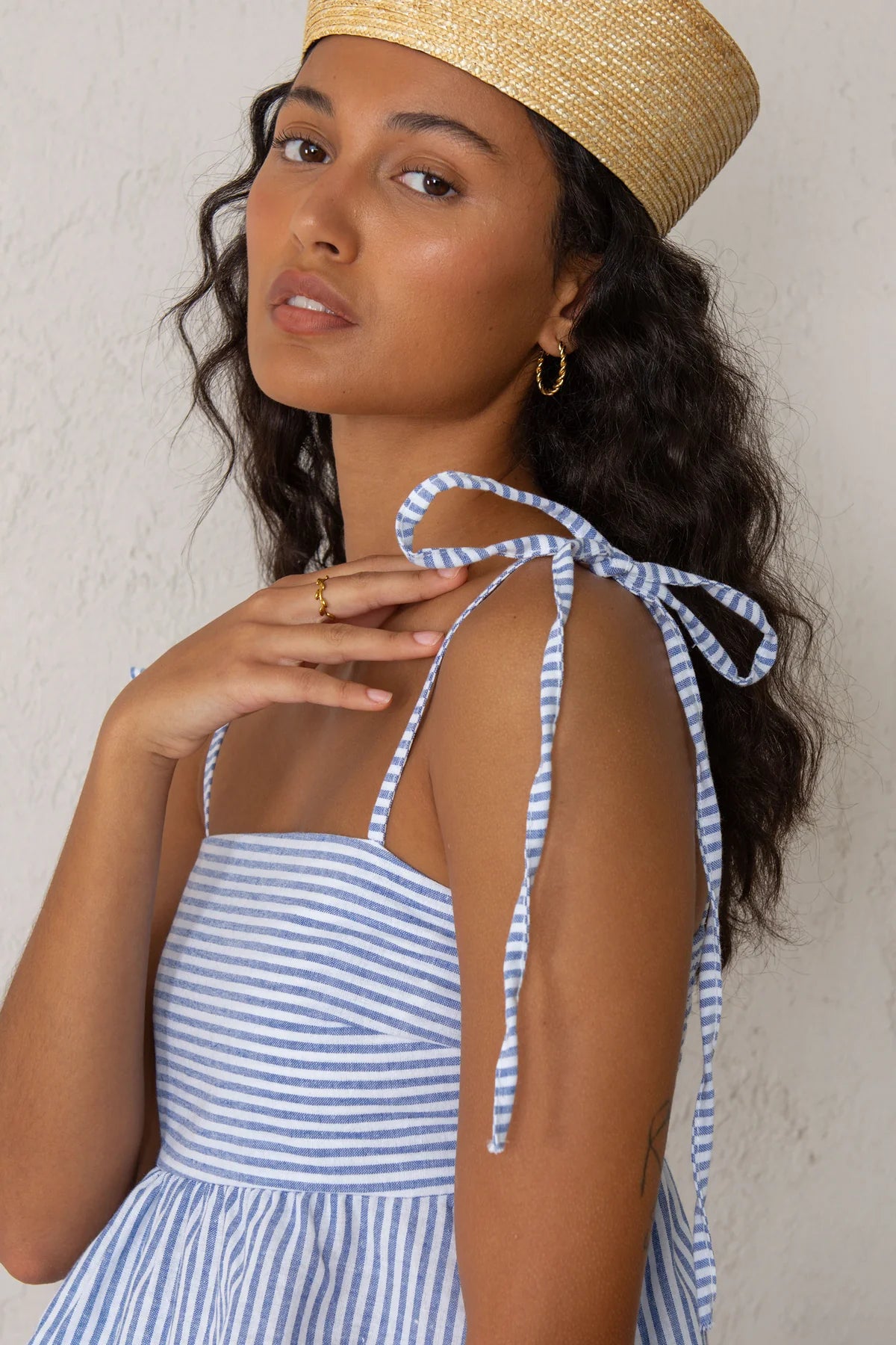 Woman wearing a blue and white striped tank top with a straw hat against a plain background
