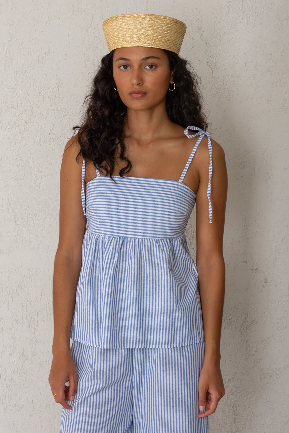 Woman wearing a blue and white striped tank top and matching pants with a straw hat against a plain background