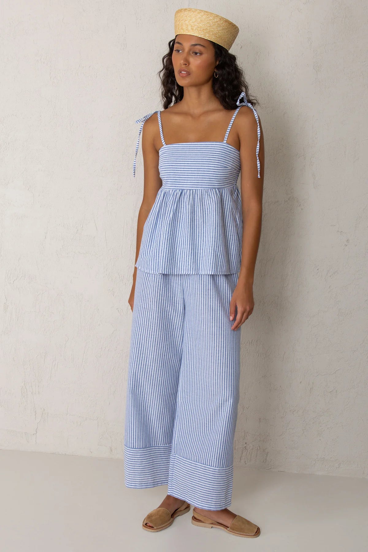 A woman modeling a light blue and white striped two-piece set consisting of a sleeveless peplum top with tie straps and matching wide-leg pants, paired with a straw hat and tan sandals against a textured off-white wall.
