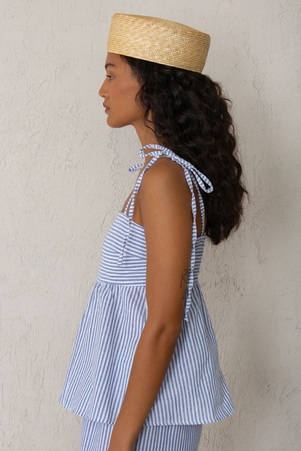 Woman wearing a blue and white striped tank top with a straw hat against a plain background