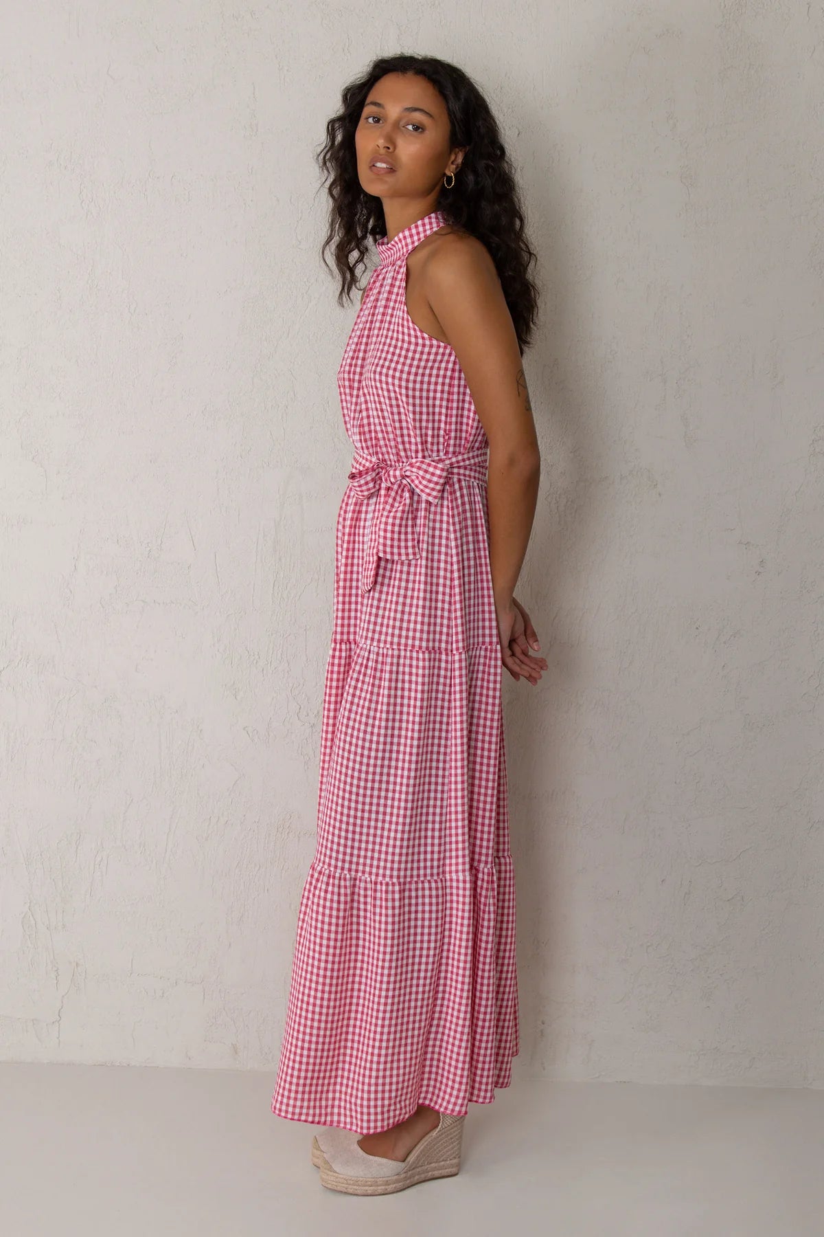 A woman wearing a pink and white gingham halter maxi dress with a tie waist, standing against a textured off-white wall.