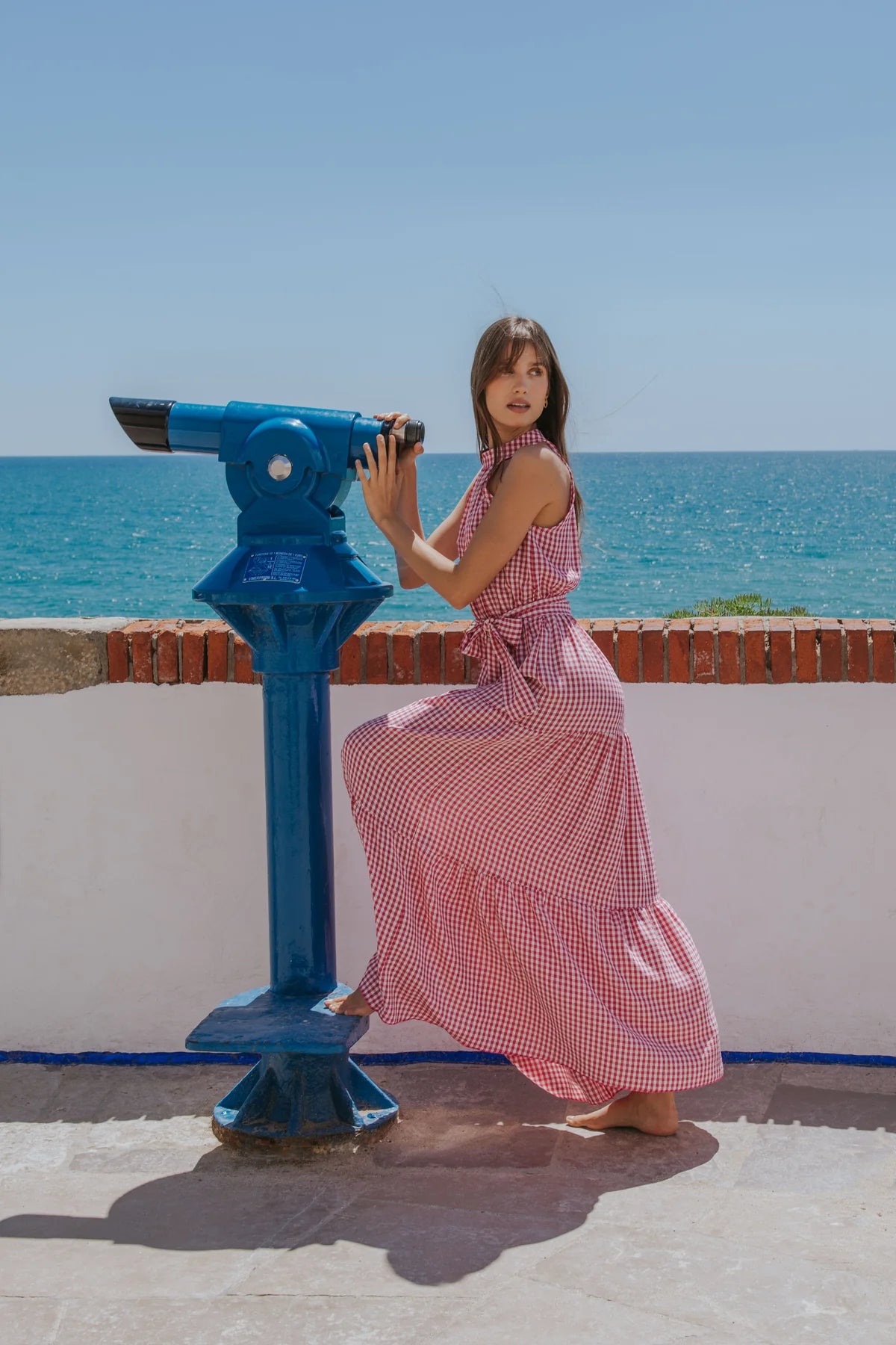 A woman in a pink and white gingham maxi dress poses next to a blue coin-operated binocular viewer on a sunny beach walkway with the blue sea and sky in the background.