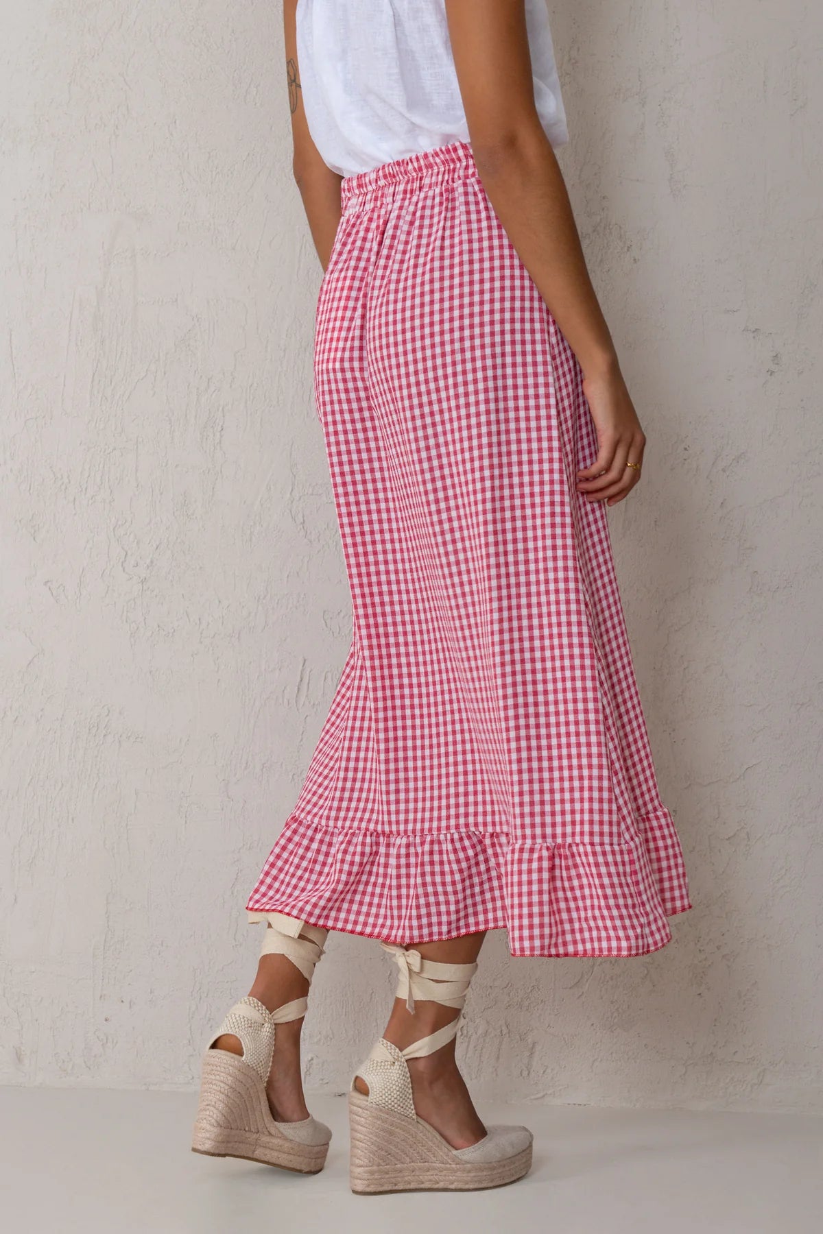 A person wearing a pink and white gingham midi skirt with a ruffled hem, paired with a white sleeveless top and beige wedge espadrille sandals, standing against a textured off-white wall.