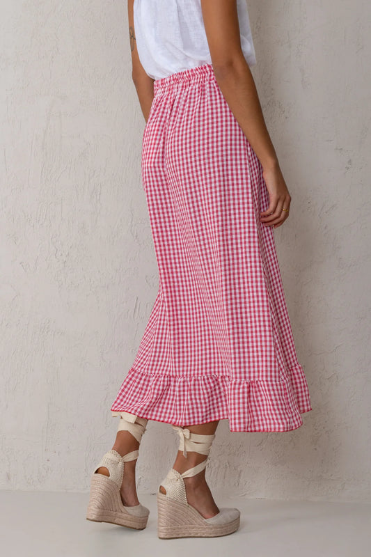 A person wearing a pink and white gingham midi skirt with a ruffled hem, paired with a white sleeveless top and beige wedge espadrille sandals, standing against a textured off-white wall.