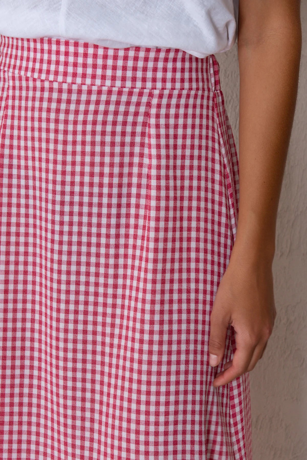 A close-up of a person wearing a red and white gingham checkered skirt with a white top, standing against a textured off-white wall.