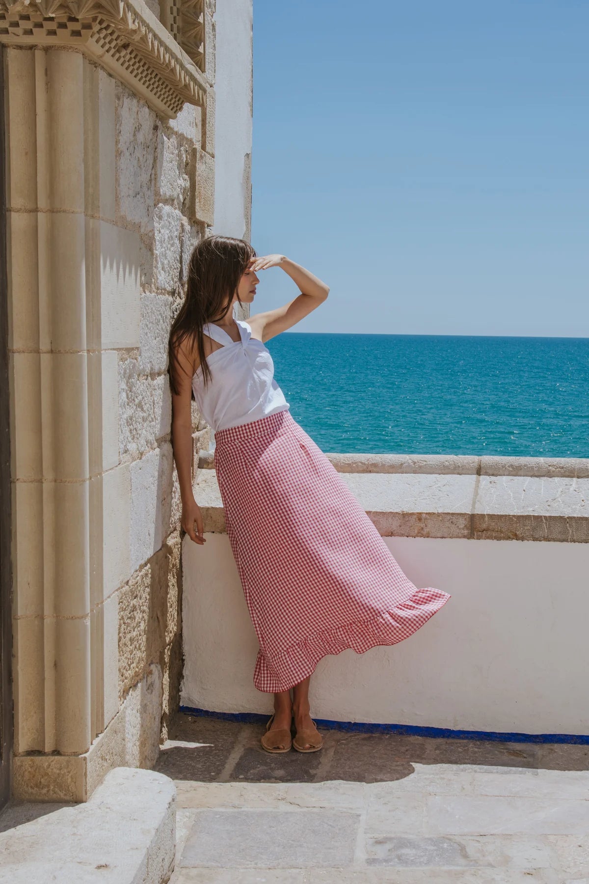 Woman in a white top and red checkered skirt standing by a stone wall with a blue sea and clear sky in the background.