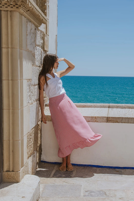 Woman in a white top and red checkered skirt standing by a stone wall with a blue sea and clear sky in the background.