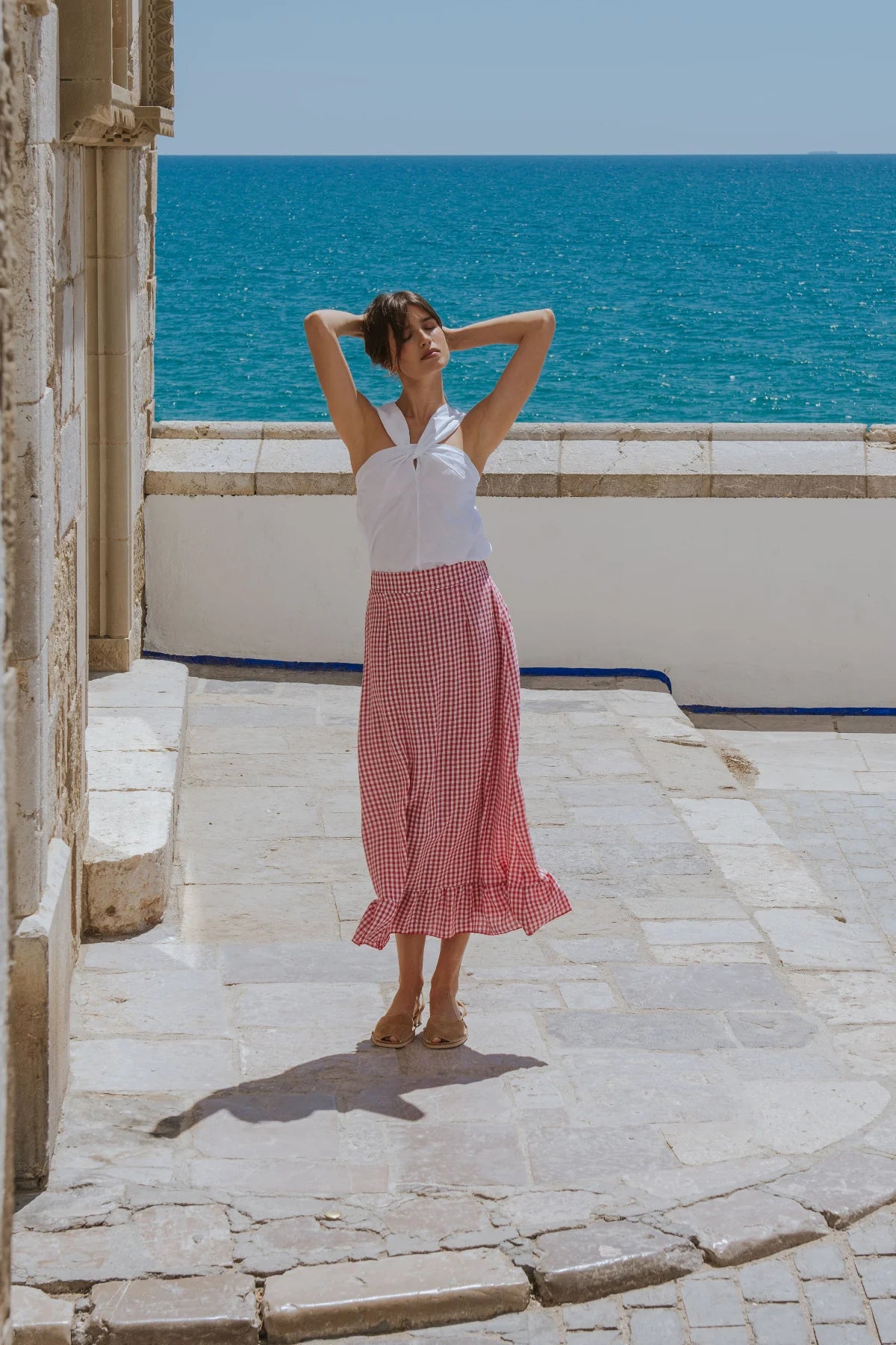 Woman in a white top and red checkered skirt standing on a stone path with ocean view.