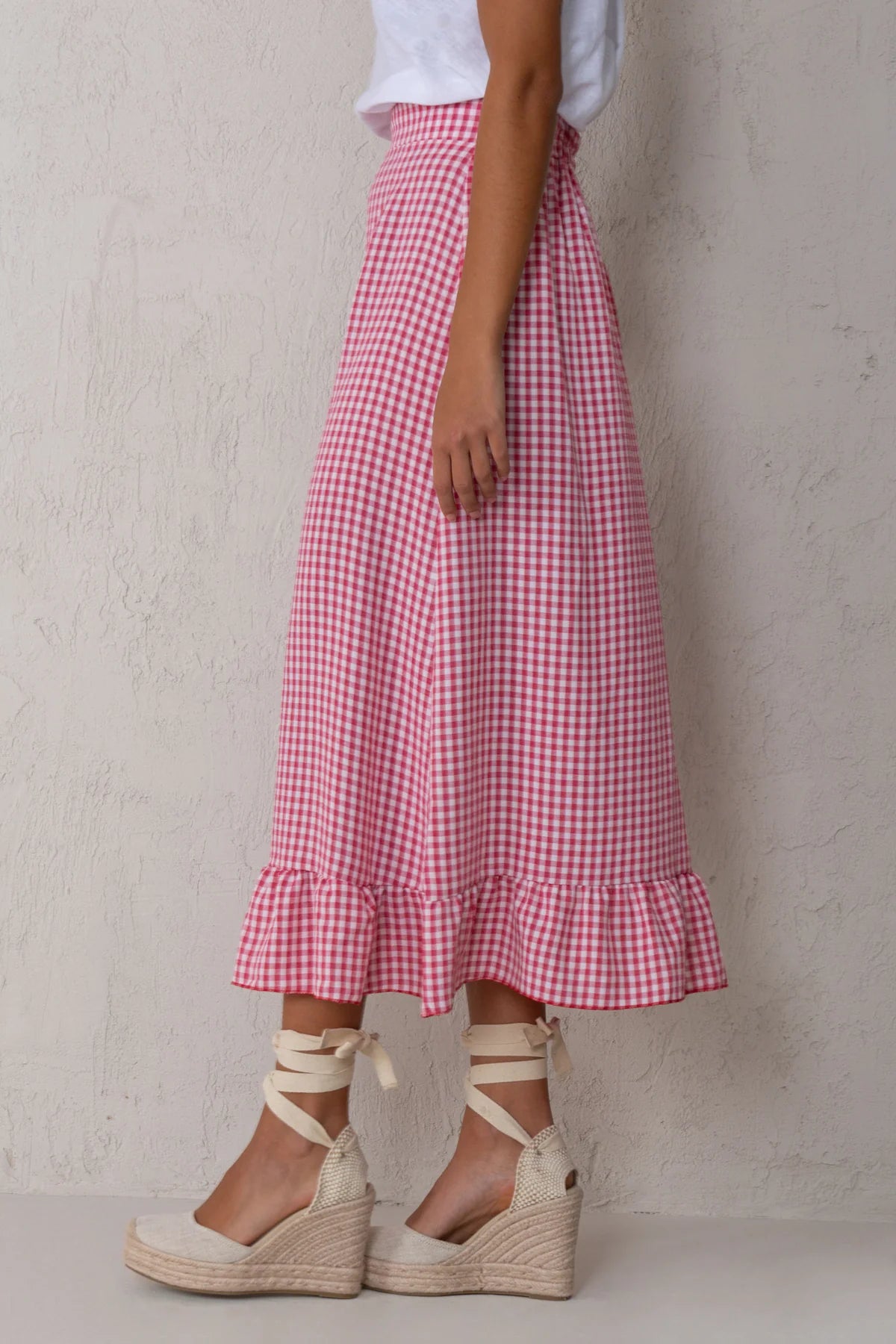A model wearing a pink and white gingham check midi skirt with a ruffled hem, styled with a white top and beige wedge espadrille sandals against a textured light gray wall.