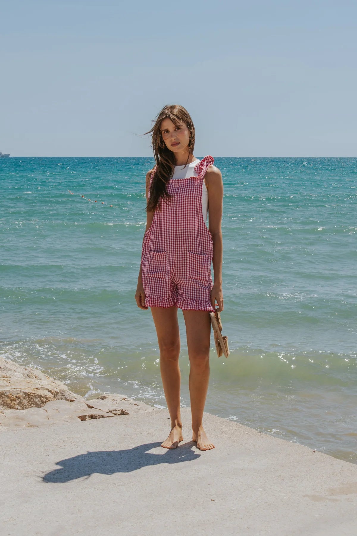A woman stands barefoot on a concrete pier by the sea, wearing a pink and white gingham overall romper with a white sleeveless top underneath. The background features a bright blue sky and turquoise ocean water.