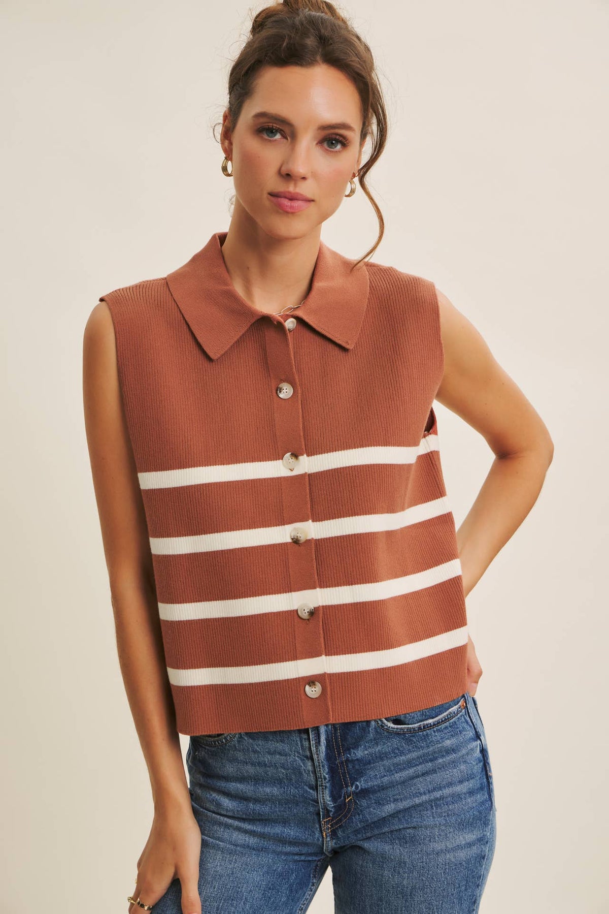 Model wearing a terracotta and cream striped sleeveless sweater vest with a collared neckline, button front, and blue jeans.