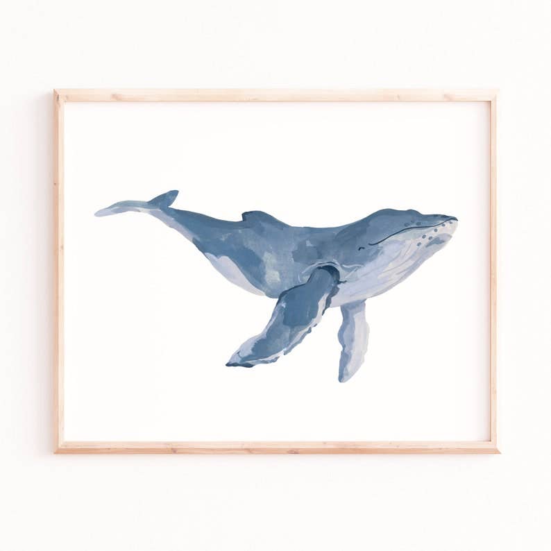 A framed watercolor art print of a blue-gray humpback whale on a white background, enclosed in a light natural wood frame and mounted on a white wall.