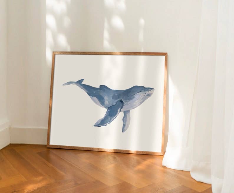 A framed whale art print leaning against a white wall on a wooden floor, with soft sunlight and curtain shadows cast across the scene.