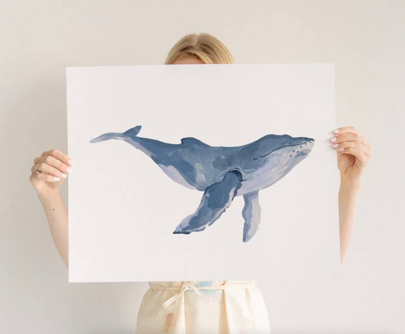 A person holding up a large white art print featuring a watercolor illustration of a blue-gray whale, shown against a plain light background.