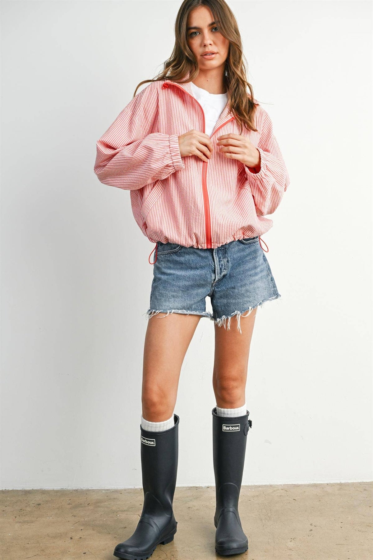 A woman modeling a light red zip-up hooded jacket, paired with denim shorts, white socks, and black rain boots, standing against a plain off-white wall.