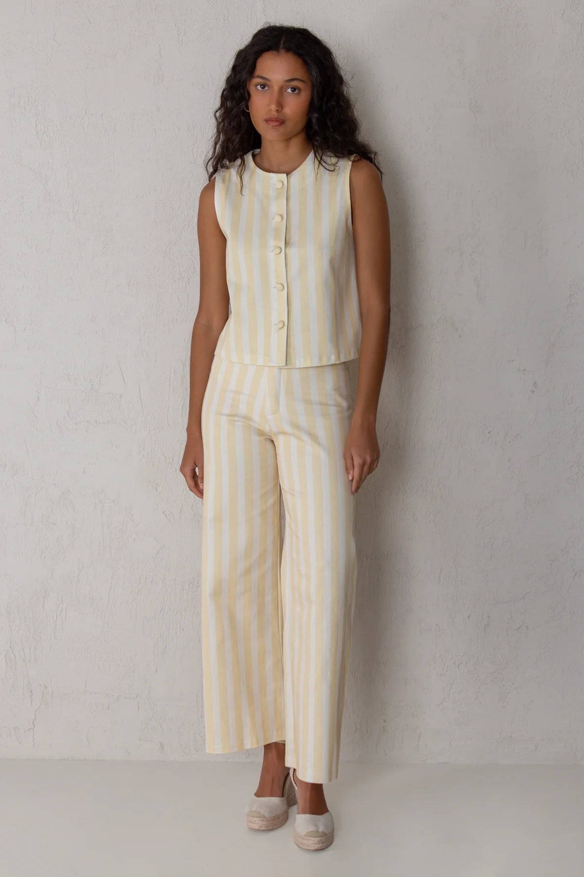A woman modeling a sleeveless button-front top and matching wide-leg cropped pants in a cream and pale yellow vertical stripe set, standing against a light textured wall.