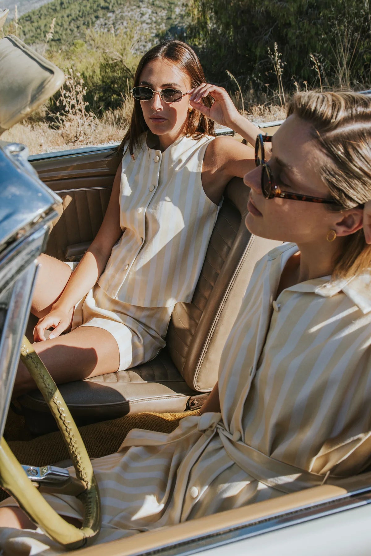 Two women wearing matching cream and tan vertically striped sleeveless dresses sit inside a vintage convertible car outdoors on a sunny day.
