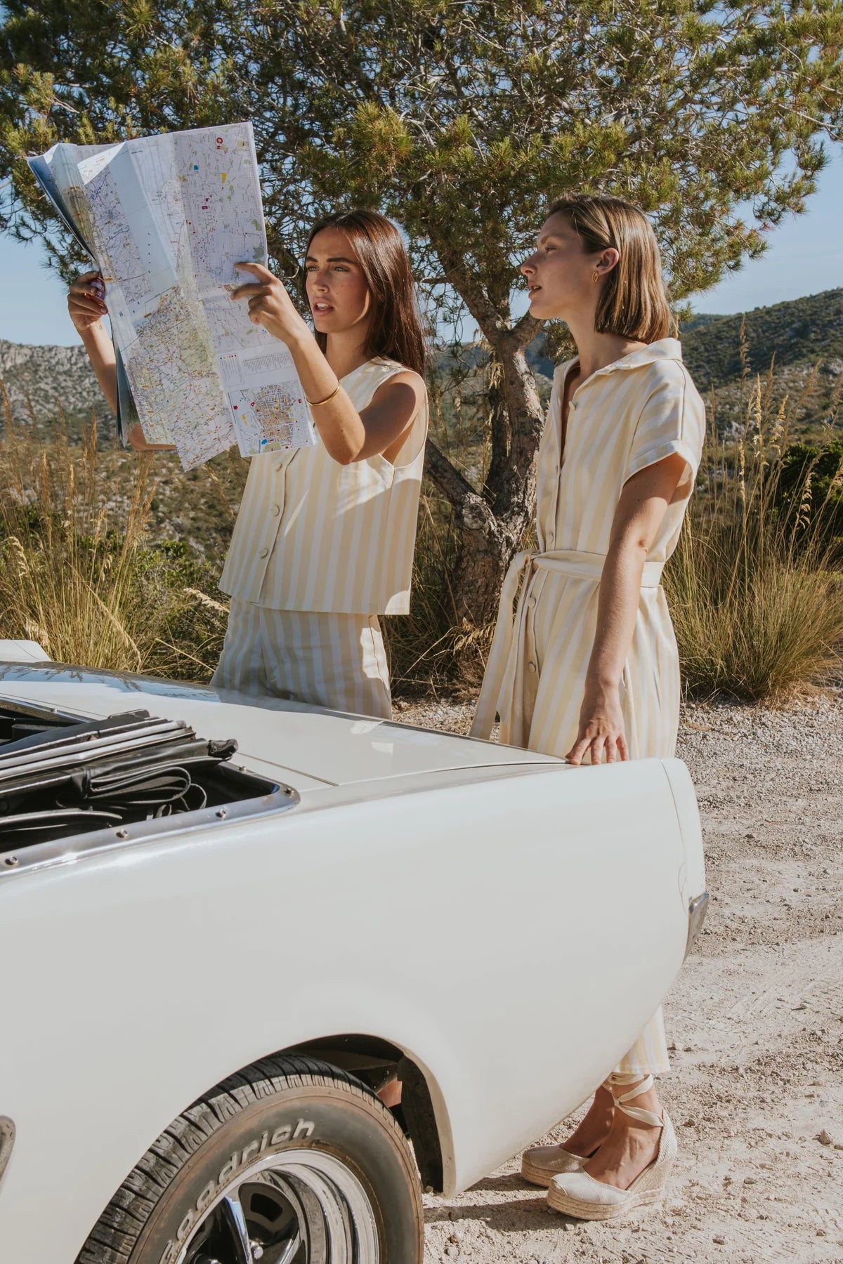 Two women standing outdoors beside a white vintage car on a dirt road, with one holding a large map. They are wearing light beige dresses, and trees and dry grass are visible in the background.