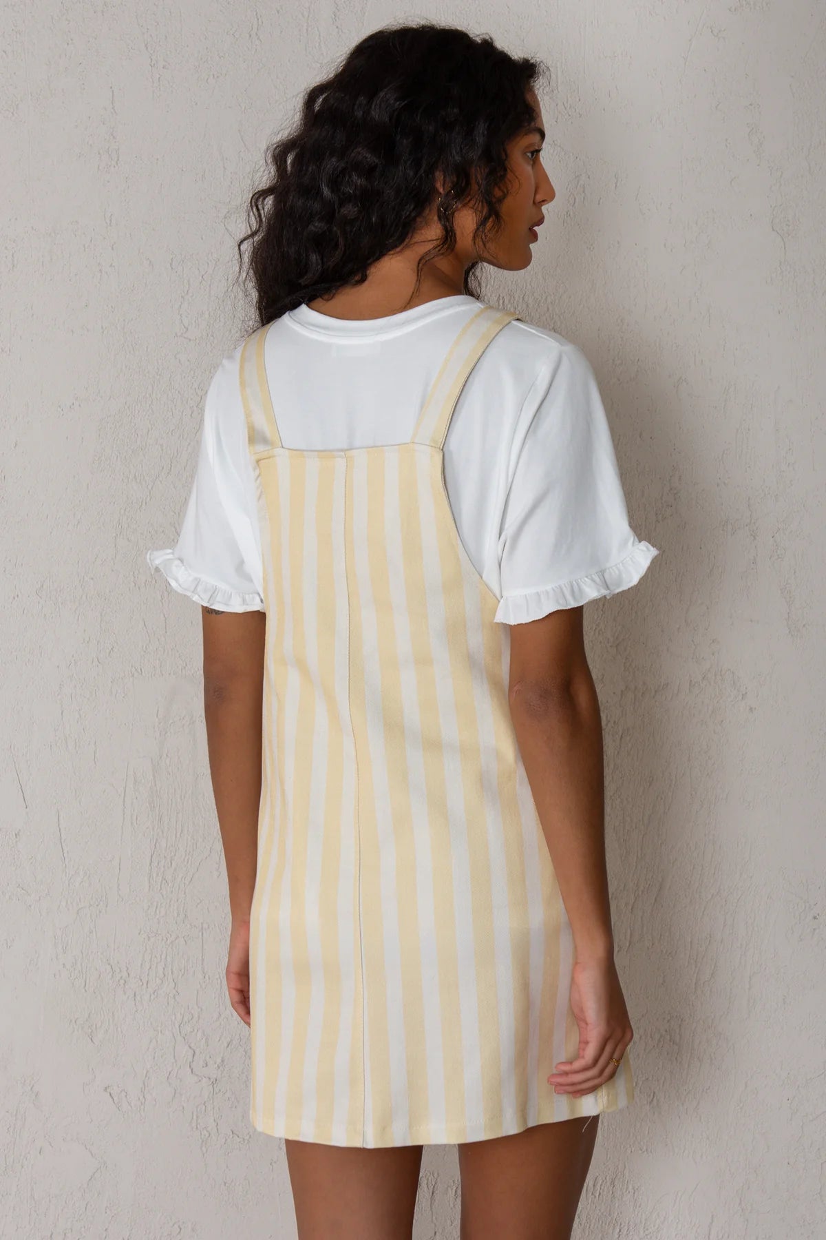 A woman shown from the back wearing a white ruffled-sleeve t-shirt layered under a pale yellow and white vertically striped pinafore dress, standing against a textured light gray wall.