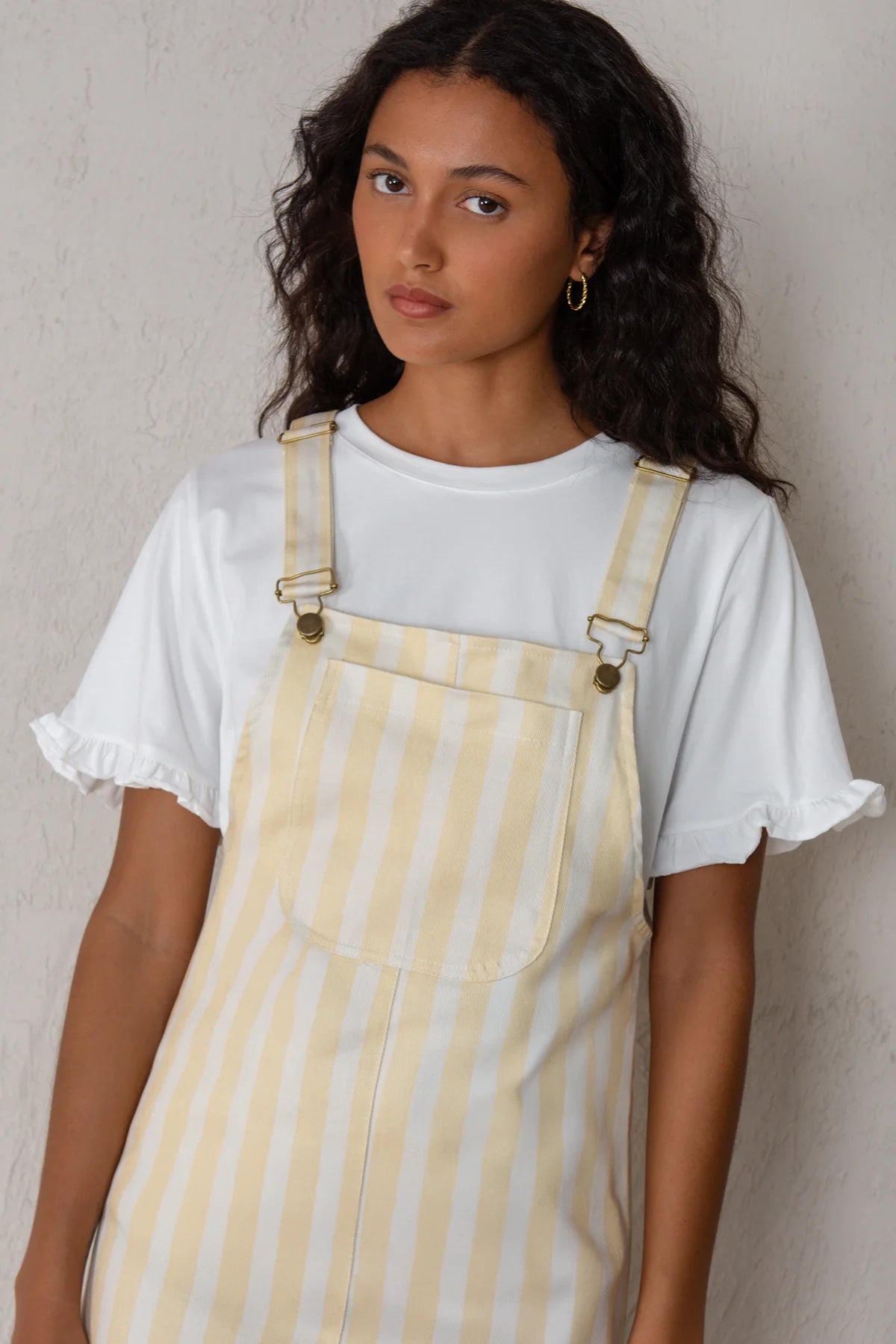 Woman wearing a white t-shirt and yellow striped overalls against a plain background