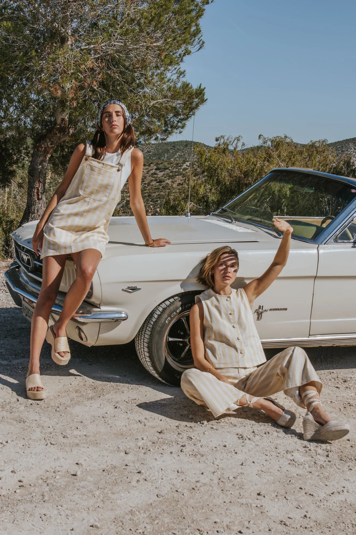 Two women in matching beige linen outfits pose with a vintage white car on a dirt road with trees in the background.