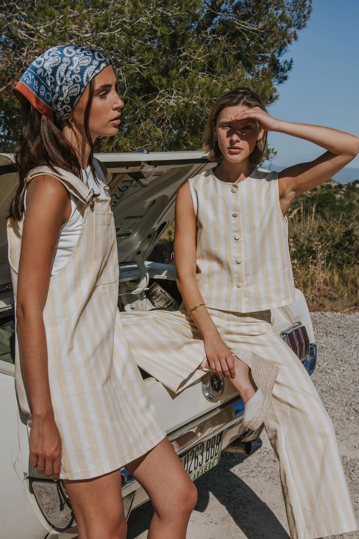 Two women wearing matching beige and white striped outfits pose in front of an open car door outdoors under a blue sky.