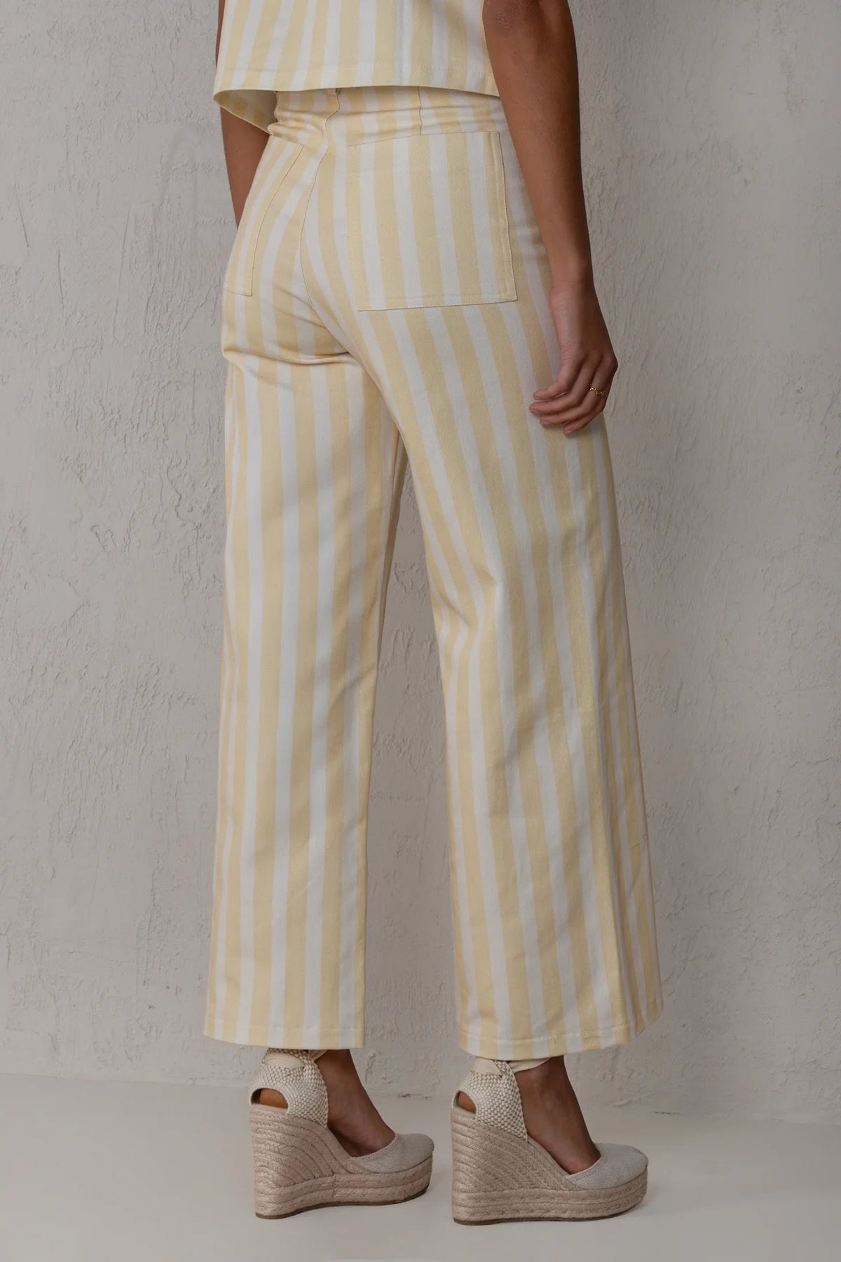 A person shown from the back wearing pale yellow and white vertically striped cropped wide-leg trousers with a matching top and beige espadrille wedge sandals, standing against a textured off-white wall.