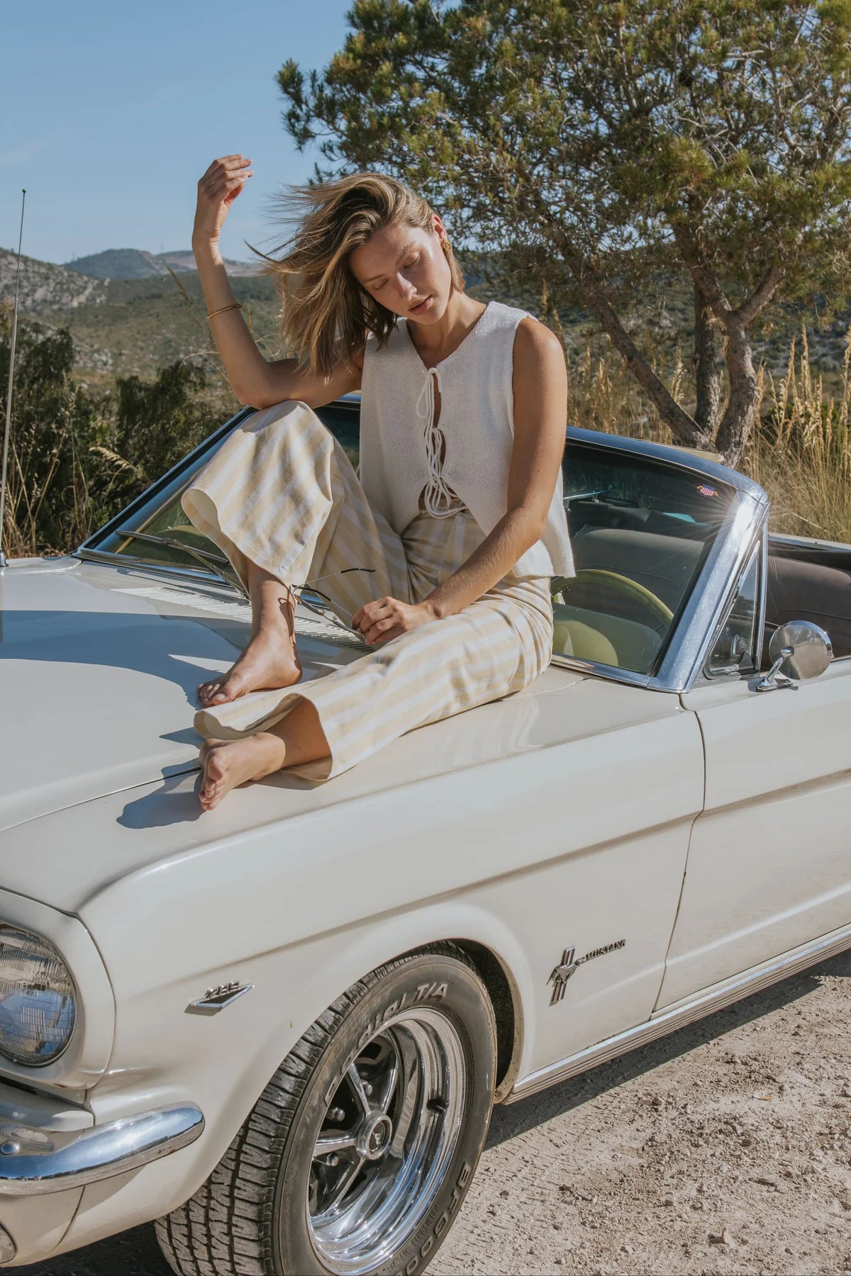A woman wearing a cream sleeveless top and matching wide-leg trousers sits barefoot on the hood of a vintage cream convertible car outdoors, with trees and hills in the background.