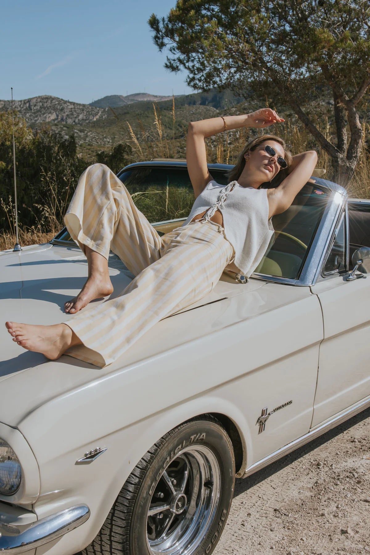 A woman wearing a white sleeveless top and beige wide-leg pants reclines on the hood of a cream-colored classic car outdoors, with mountains, trees, and a dirt road in the background.