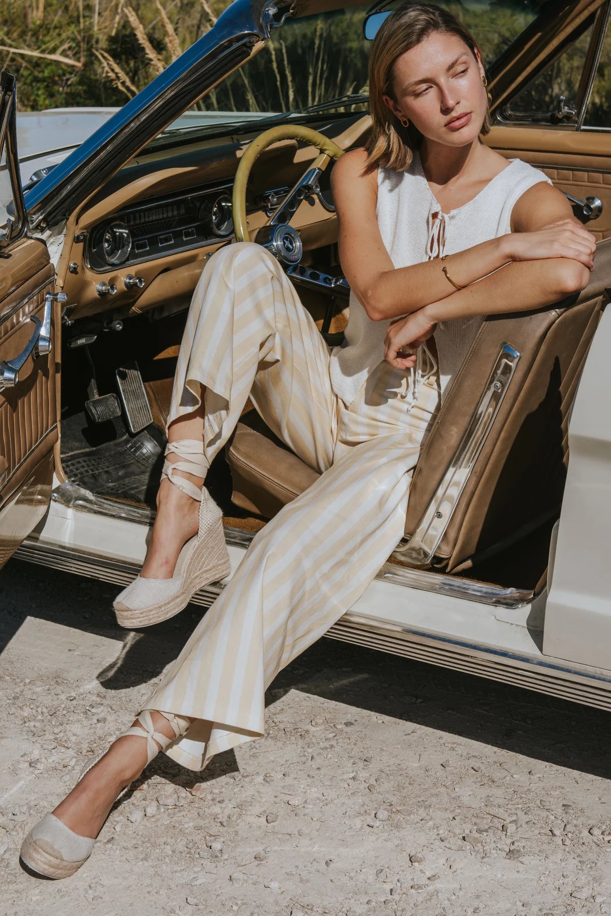 A woman wearing a cream and beige striped sleeveless top and matching wide-leg trousers sits in the open door of a vintage car, wearing beige wedge sandals.