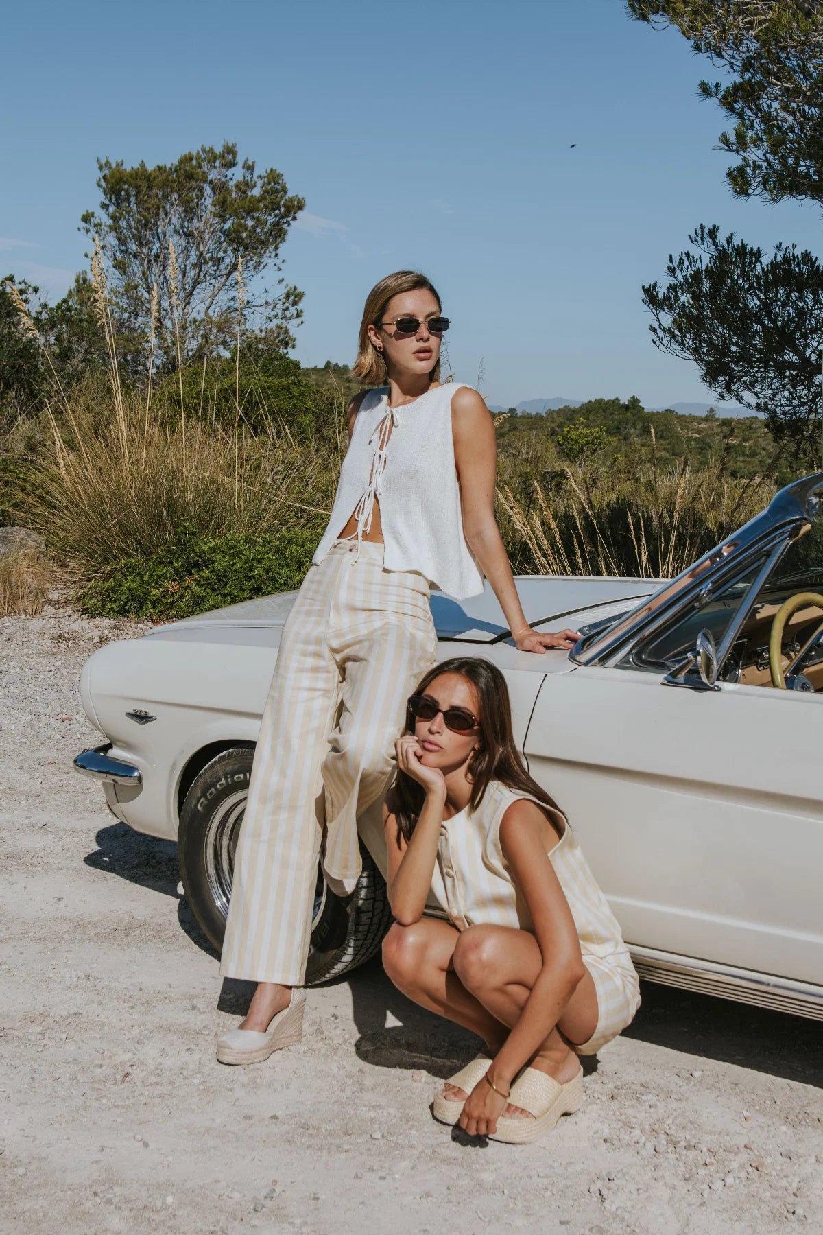 Two women posing outdoors next to a vintage white convertible car on a sandy ground under a clear blue sky. Both are wearing matching sleeveless white outfits with light beige striped pants or shorts and beige flat sandals.