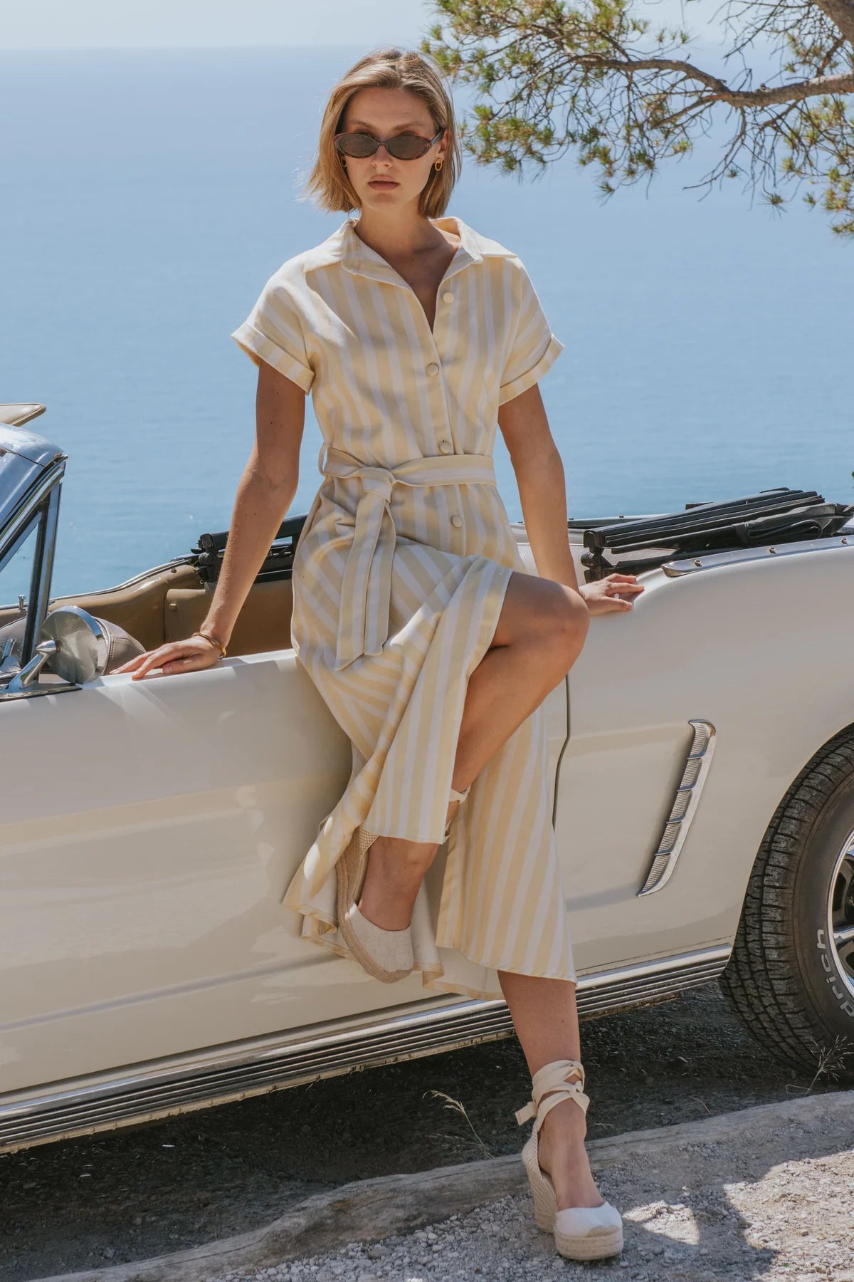 A woman wearing a cream and beige vertically striped shirt dress with a tie waist, sitting on the side of a white vintage convertible car with the sea in the background.