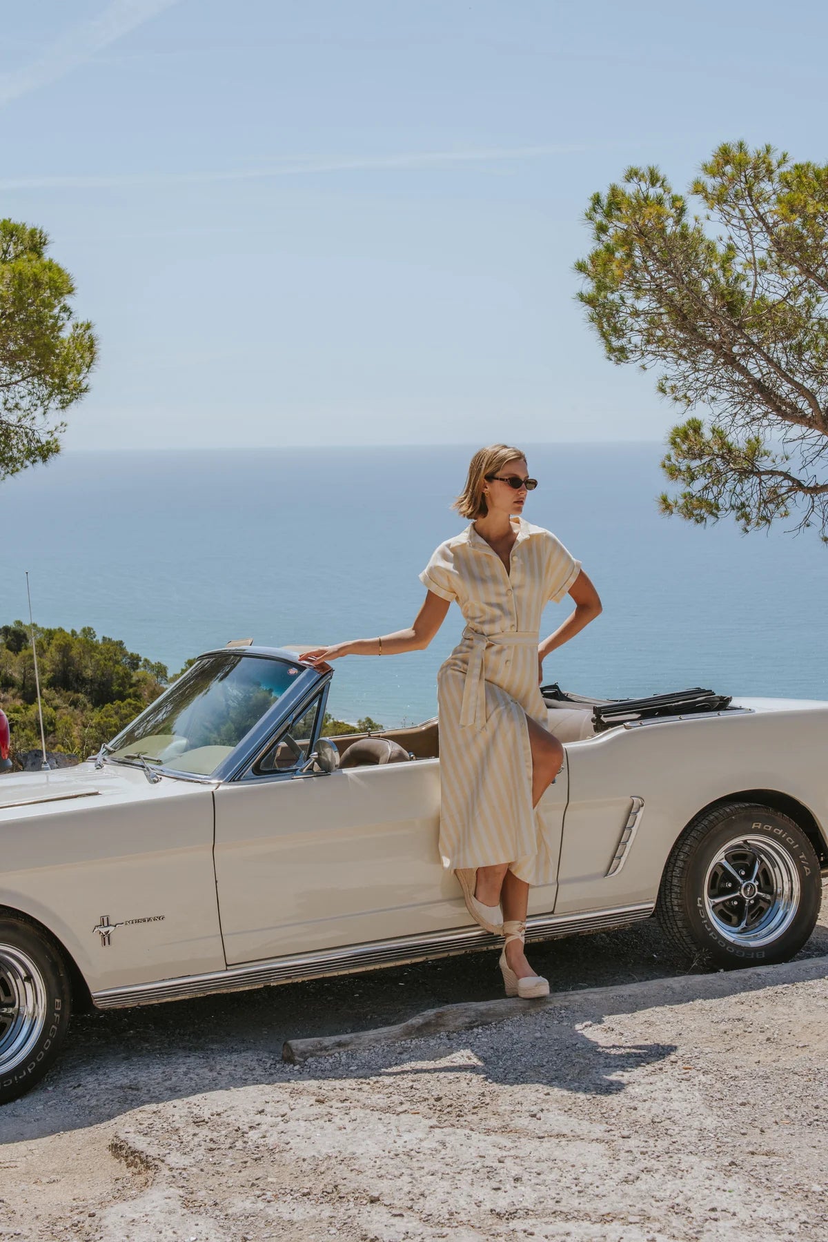 A woman in a cream-colored pleated dress and sunglasses stands next to a vintage cream convertible car on a rocky coastal path with the sea and sky in the background.